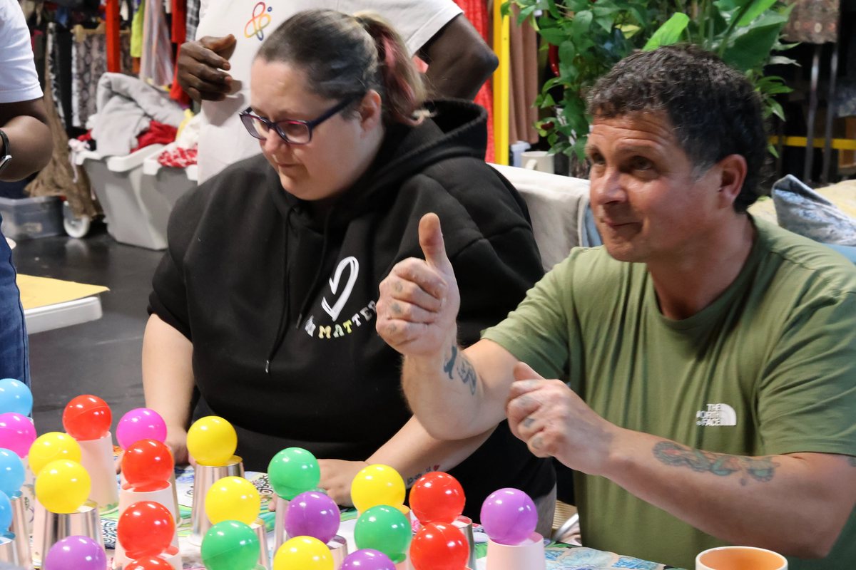 Two adults at a table scattered with brightly coloured balls, one of them giving a thumbs up.