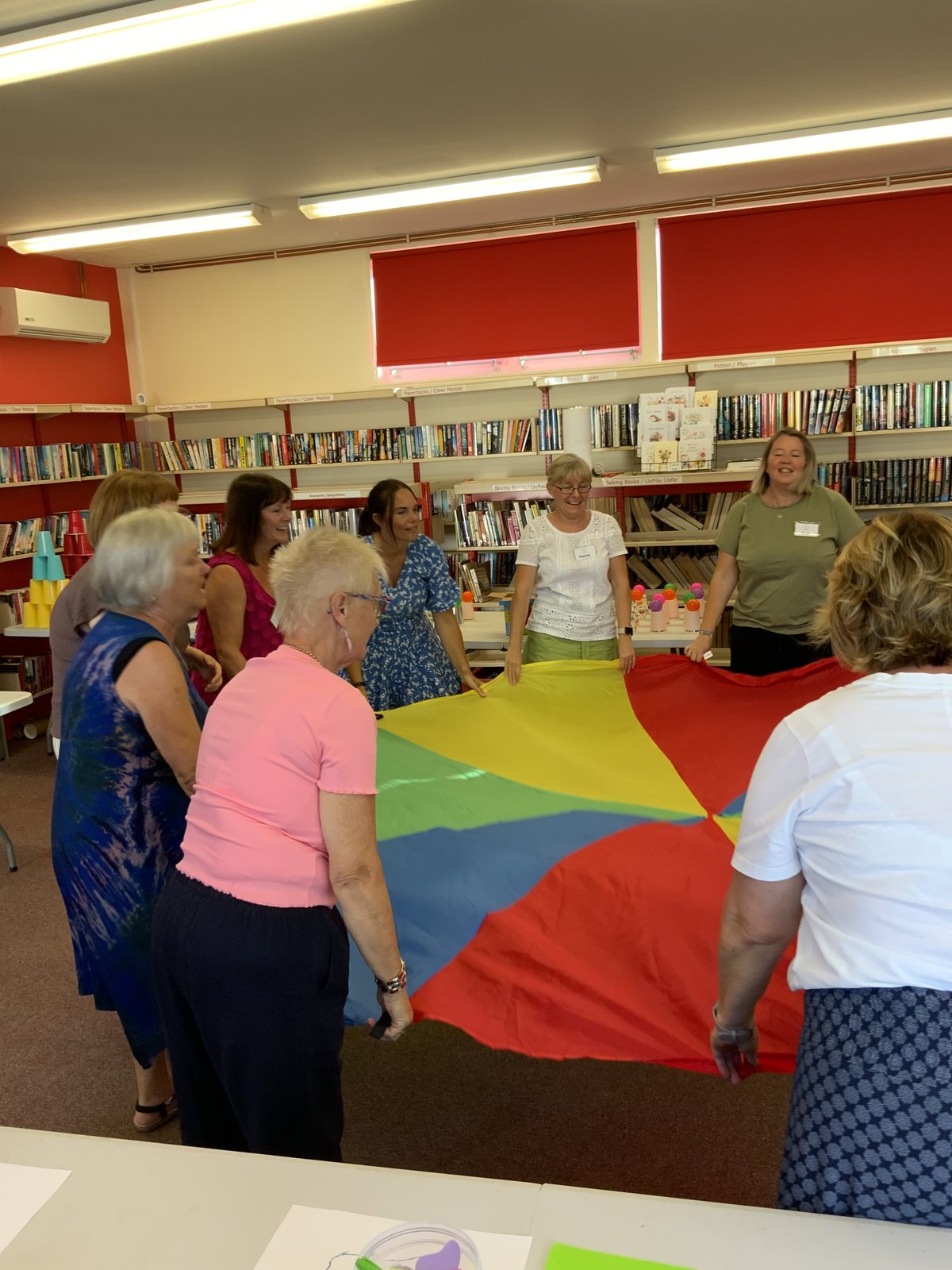 Adults gathered around a brightly coloured parachute spread out between them in a library, with others watching from behind.