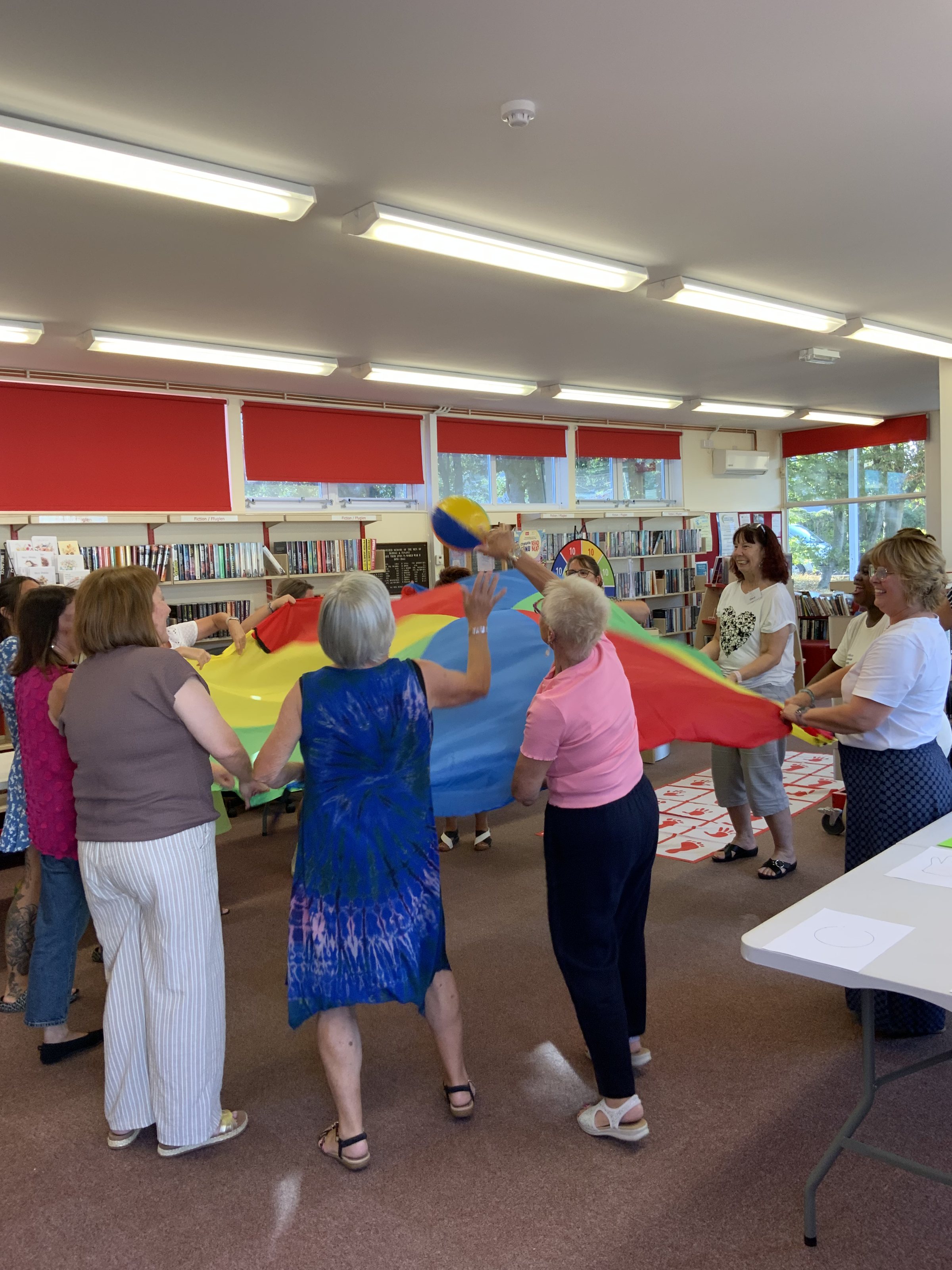 A group of adults standing in a circle in a library, lifting a red, blue and yellow parachute above their heads.