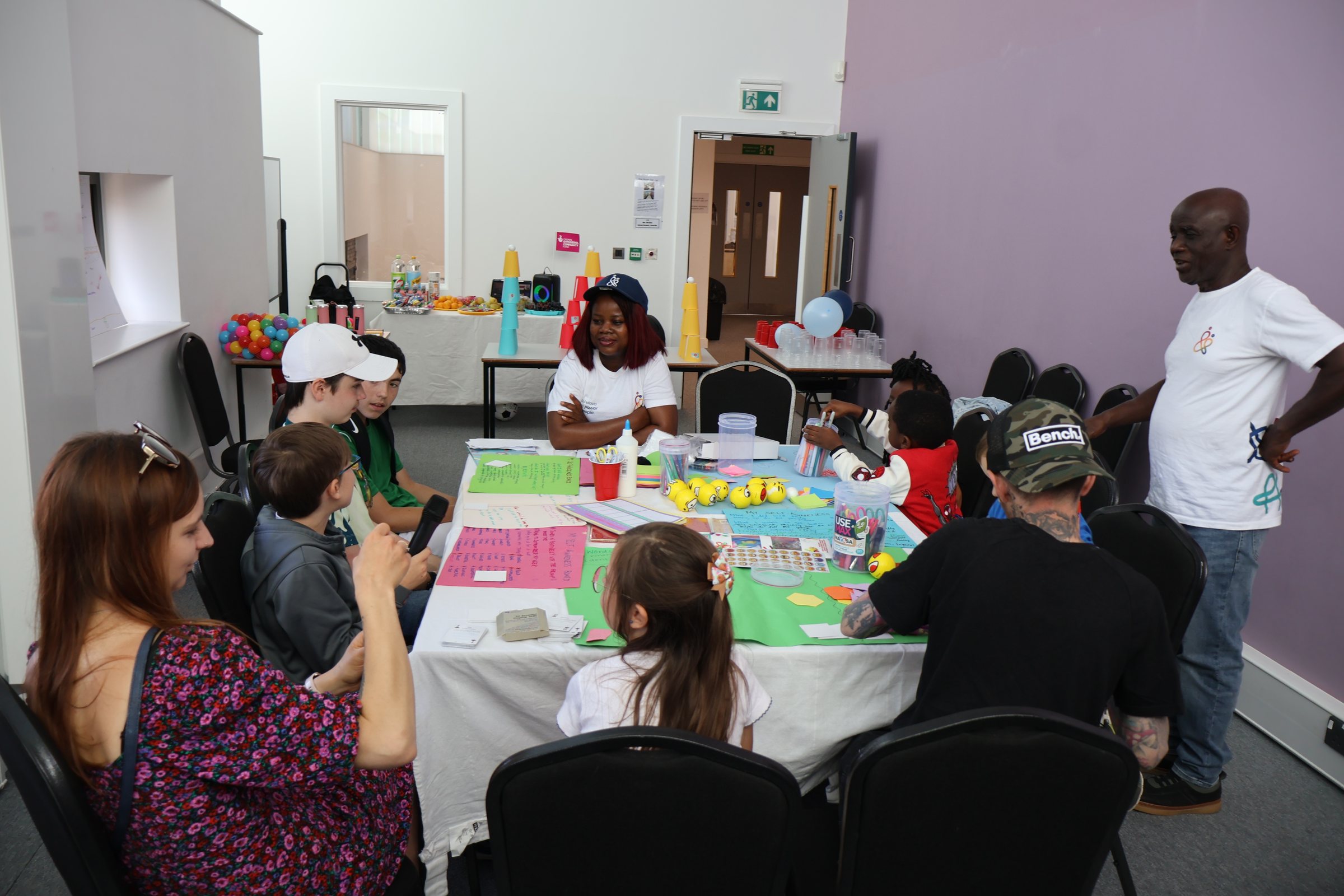 A wide view of a full session: two long tables of young people and facilitators working with craft materials in a community room.