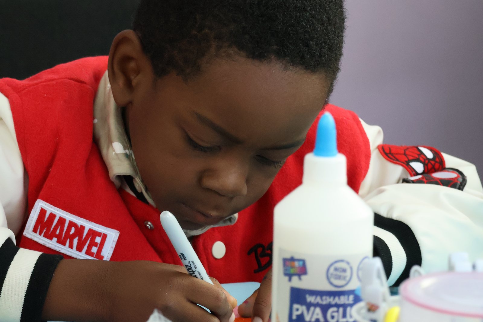 A boy in a red jacket writing carefully on a piece of card, glue and pens beside him.