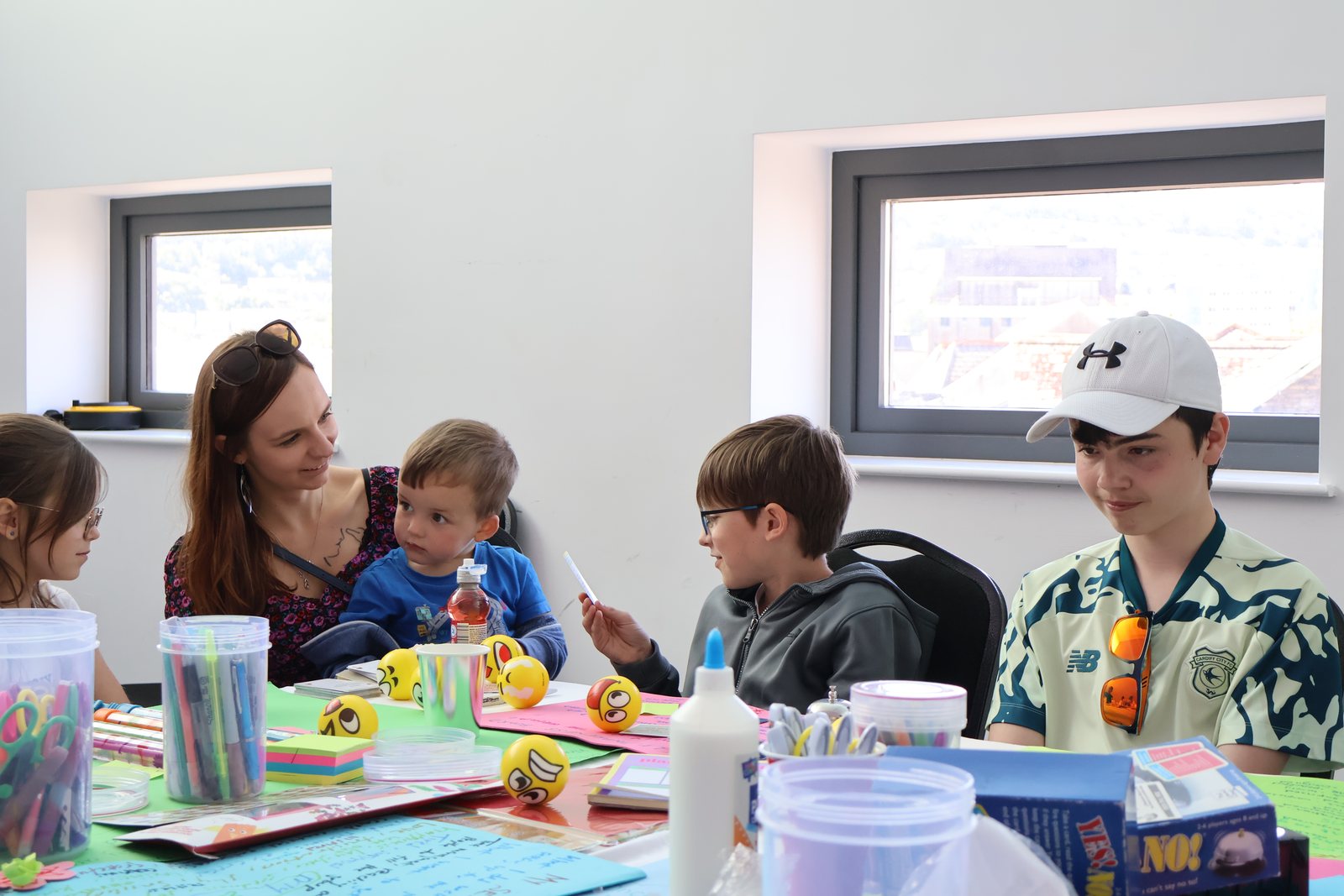 A group of adults and children around a craft table, including a small child on an adult's lap, all working with paints and paper.