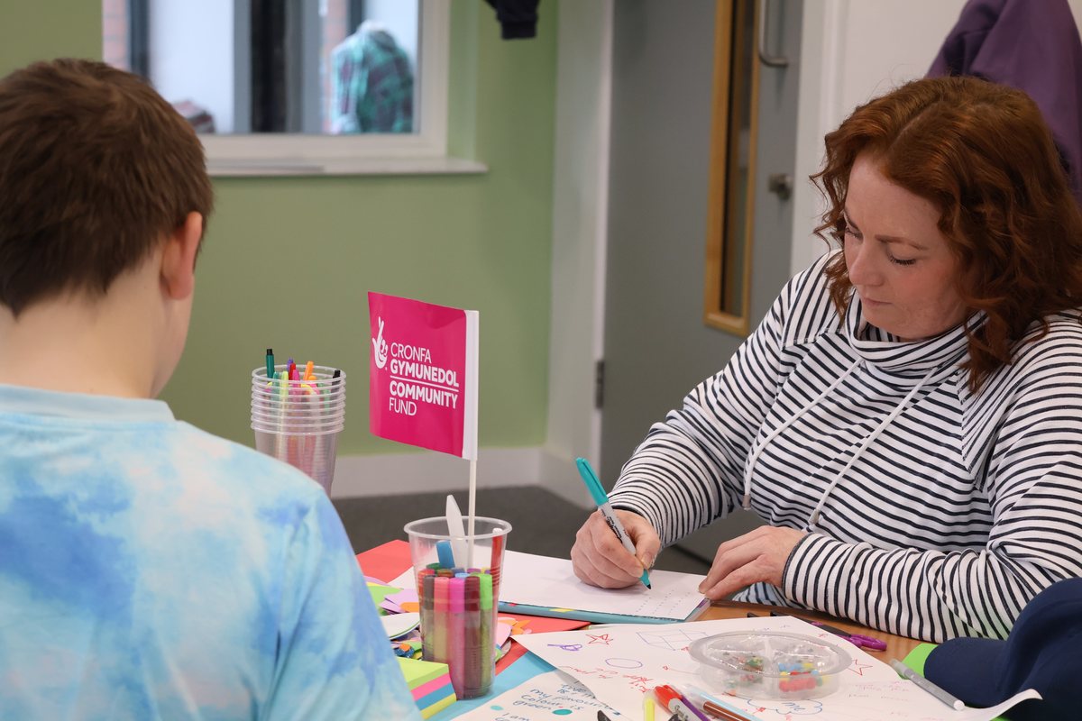 An adult writing at a craft table beside a Cronfa Gymunedol Community Fund flag.