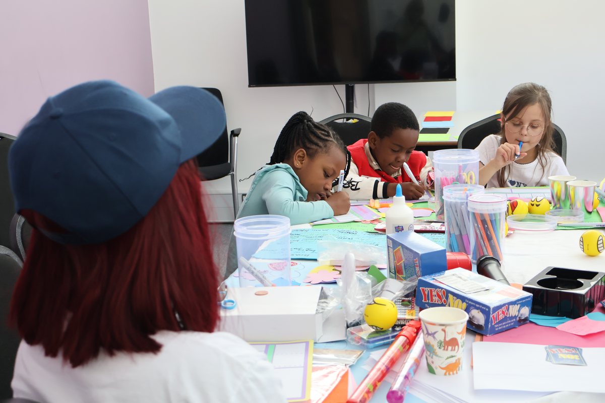 A busy craft table with several young people working, materials spread across it.