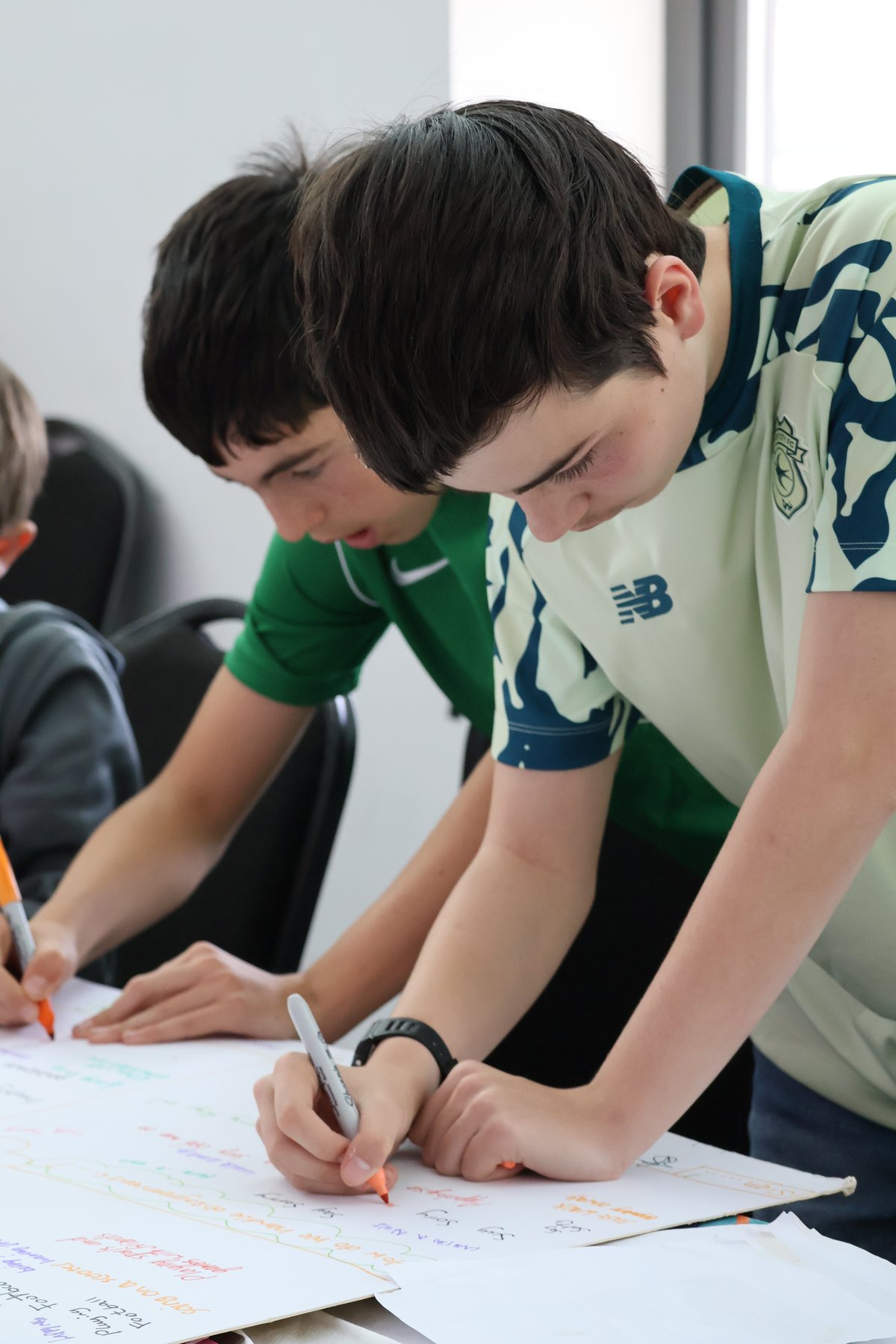 Close-up of two young people writing on a large sheet of paper on an easel, an adult's hand steadying it.