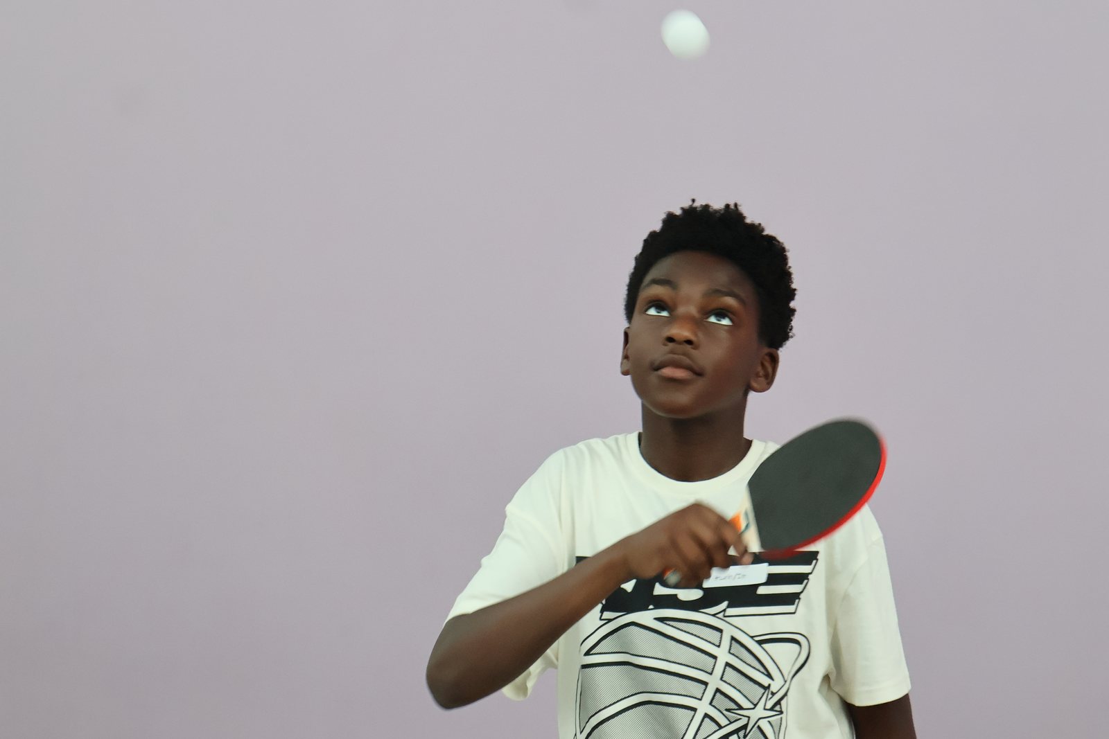 A boy mid-serve with a table tennis bat, the ball in the air above him.