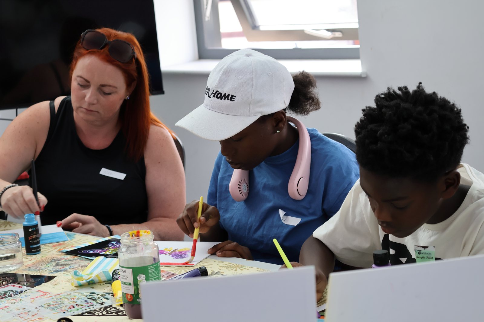 A facilitator sitting between two young people, helping them paint at a tabletop easel.