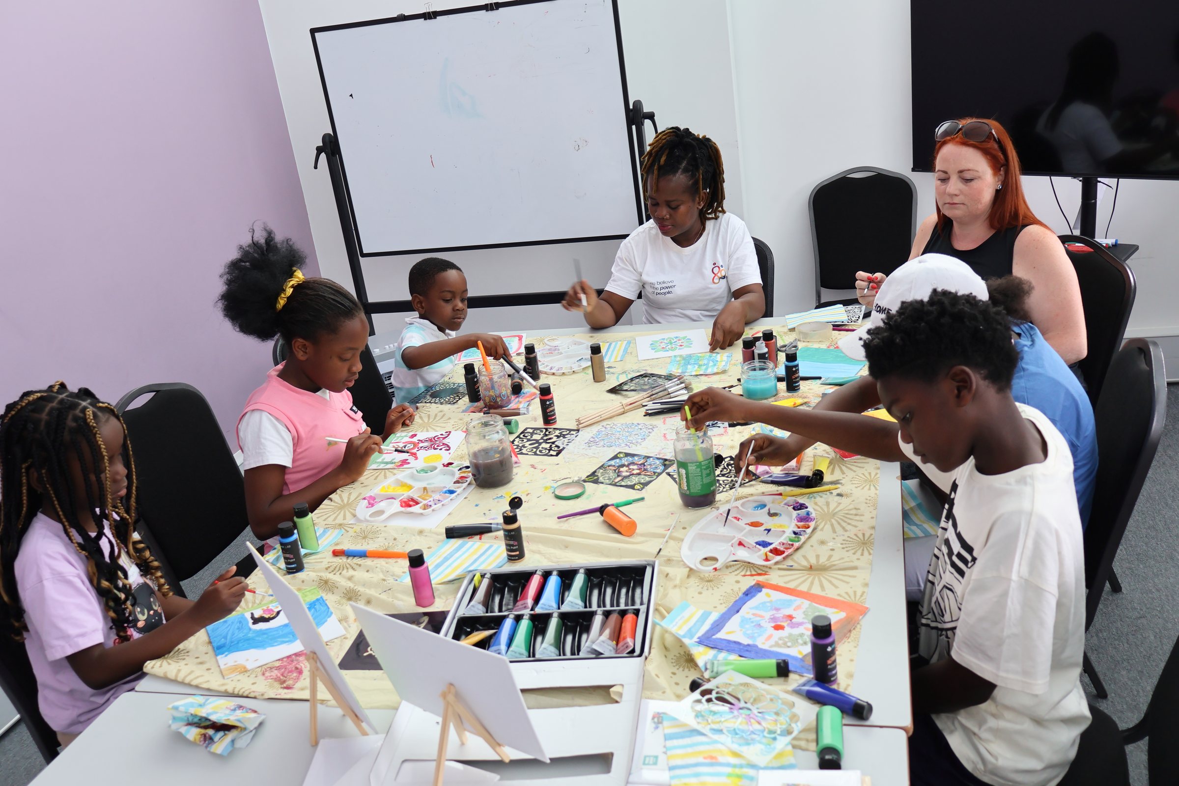 A busy craft session in a bright room: a dozen young people and facilitators at a long table strewn with paints, pens and finished artwork on small easels.