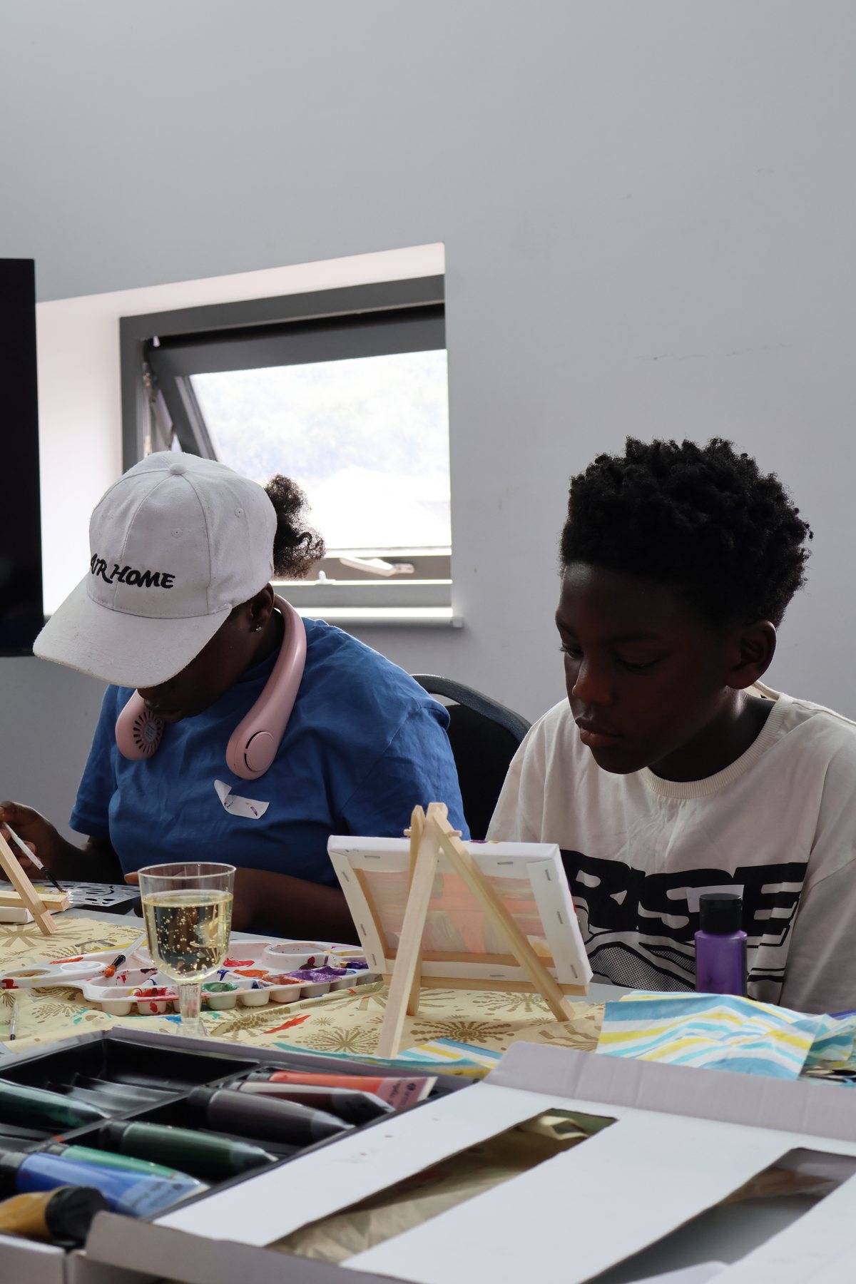 Two young people painting at a small tabletop easel, brushes and paint pots in front of them.